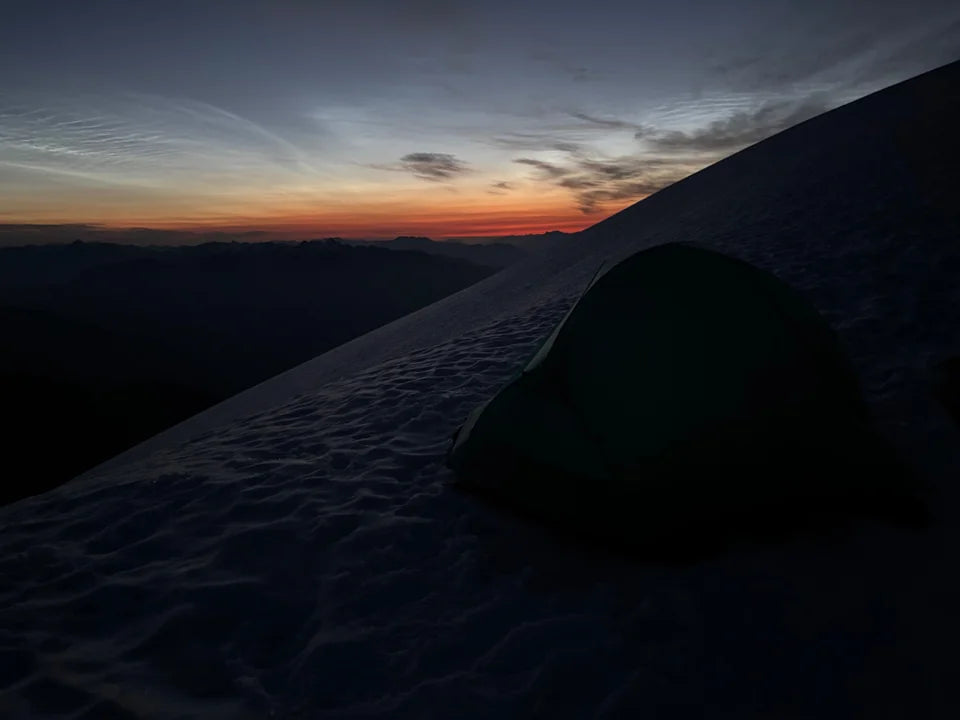 A tent on snow silhouetted against sunrise over distant mountains